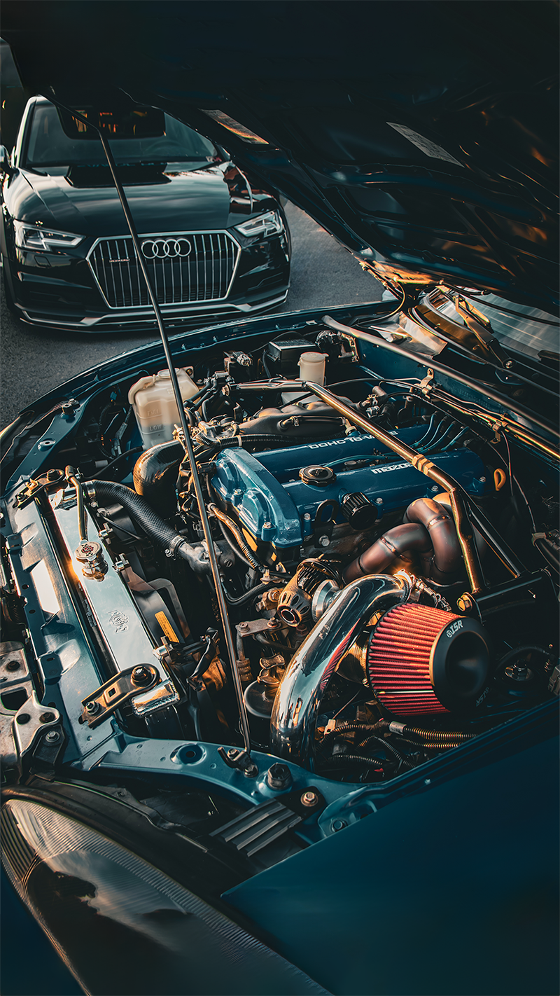 Photo of the engine bay of a blue 2001 Mazda MX-5 Miata NB with an audi in the background